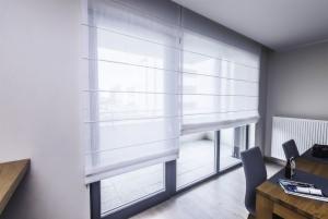 Jasne, białe rolety rzymskie zamontowane w oknie balkonowym, widok z wnętrza nowoczesnego salonu z drewnianym stołem i szarymi krzesłami. Delikatne światło dzienne podkreśla fakturę materiału rolet.