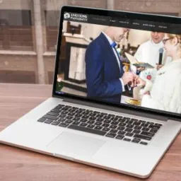 Laptop wyświetla zdjęcie ślubne pary podczas ceremonii zakładania obrączek, z logo 'Emotions Photography' w lewym górnym rogu ekranu, umieszczony na drewnianym biurku.