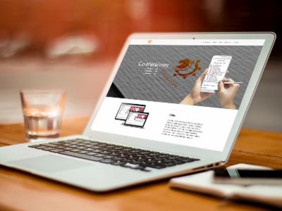 Laptop wyświetlający stronę internetową z grafiką konferencji, obok szklanka wody i notatnik z długopisem na drewnianym biurku.