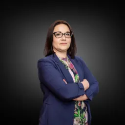 Portret kobiety w okularach i niebieskiej marynarce, z założonymi rękami na tle gradientu szarości, reprezentującej prawnika z Pasym.