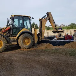 Żółta koparko-ładowarka CAT zasypuje fundamenty domu jednorodzinnego, widoczni pracownicy w odzieży roboczej, w tle domy w zabudowie jednorodzinnej.