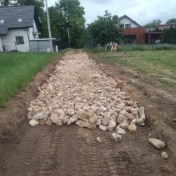 Przygotowanie podłoża pod drogę: wykop wypełniony grubym kruszywem na tle domów jednorodzinnych i zieleni.