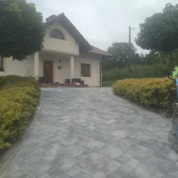 Nowo ułożony, szary podjazd z kostki brukowej prowadzący do domu jednorodzinnego, z zagęszczarką w tle i ozdobnymi krzewami po bokach.