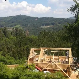 Surowy stan domu z bali w trakcie budowy, widok z góry na tle zalesionych wzgórz i błękitnego nieba z chmurami.
