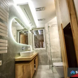 Nowoczesna łazienka z szarą marmurową okładziną, podświetlanym owalnym lustrem, drewnianą szafką pod umywalką i kabiną prysznicową ze szklanymi drzwiami.