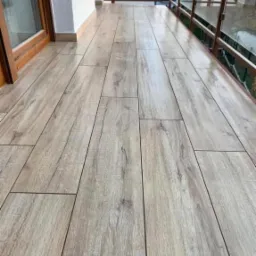 Jasne, drewnopodobne płytki ułożone na podłodze balkonu, widok z perspektywy, balustrada z metalowymi elementami i szklanymi panelami w tle.