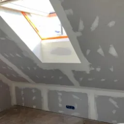 Poddasze w trakcie prac wykończeniowych: płyty karton-gips na skosach i ścianach, szpachlowane łączenia, okno dachowe z pomarańczową ramą, widoczne narzędzia pracy i wiadro z masą szpachlową...