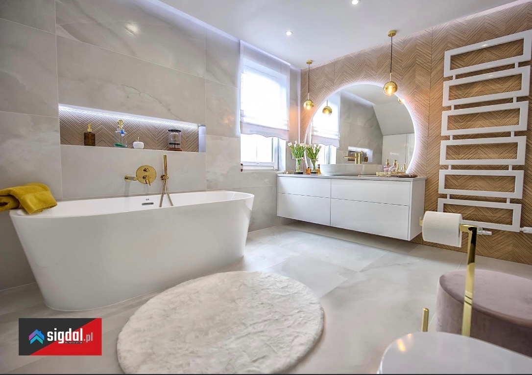 Nowoczesna łazienka w jasnych barwach z wolnostojącą wanną, okrągłym lustrem z podświetleniem LED i złotymi akcentami, takimi jak bateria wannowa i uchwyt na papier toaletowy.