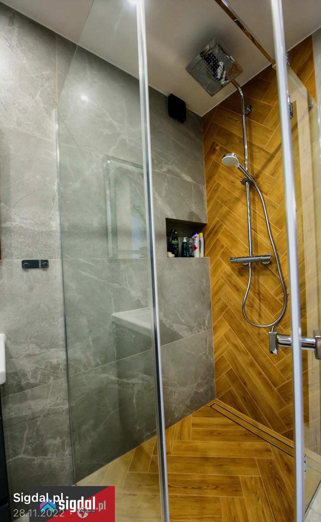 Nowoczesna kabina prysznicowa z szarymi marmurkowymi płytkami i drewnopodobną ścianą w jodełkę, wbudowana półka z kosmetykami.