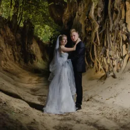 Para młoda w plenerze: panna młoda w białej sukni i welonie, pan młody w czarnym garniturze, pozują w malowniczym wąwozie z odsłoniętymi korzeniami drzew i piaszczystymi ścianami.