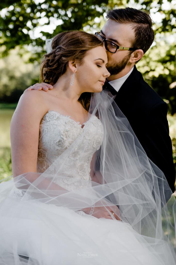 Panna młoda w sukni ślubnej z gorsetem w róże, z welonem i delikatną biżuterią, obejmowana przez pana młodego w czarnym smokingu i muszce, który całuje ją w skroń, na tle zieleni.