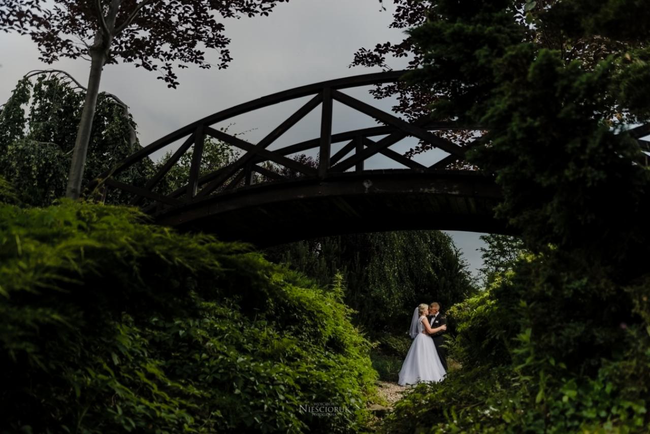 Para młoda w objęciach pod drewnianym mostkiem w otoczeniu bujnej zieleni, pochmurne niebo w tle.