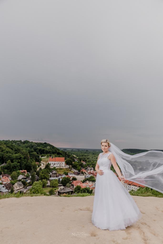 Panna młoda w białej sukni i welonie na wzgórzu z widokiem na miasto i kościół w pochmurny dzień.