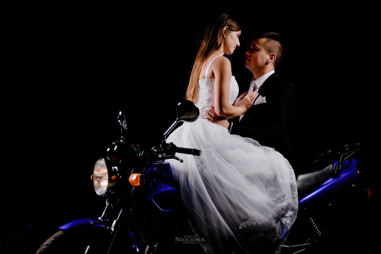 Para młoda w strojach ślubnych siedzi na niebieskim motocyklu na czarnym tle, panna młoda spogląda na pana młodego
