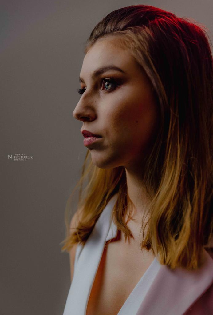 Portret profilowy młodej kobiety z delikatnym makijażem, oświetlonej czerwonym światłem z góry i boku, z widocznym logo fotografa w lewym dolnym rogu.