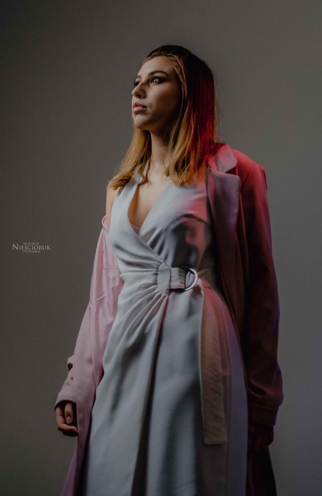Portret kobiety w jasnej sukience z drapowaniem i różowym płaszczu, oświetlonej czerwonym światłem z boku, spoglądającej w górę.