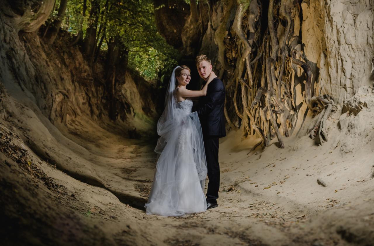 Para młoda w plenerze: panna młoda w białej sukni i welonie, pan młody w czarnym garniturze, pozują w malowniczym wąwozie z odsłoniętymi korzeniami drzew i piaszczystymi ścianami.