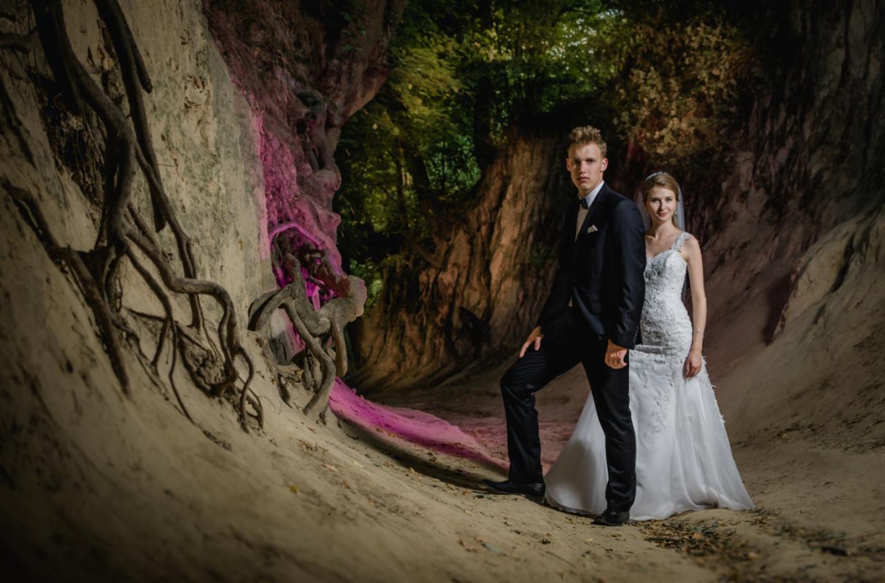 Para młoda w strojach ślubnych pozuje w nietypowej scenerii: piaszczysta grota z korzeniami i zielonym tłem, oświetlona różowym światłem.