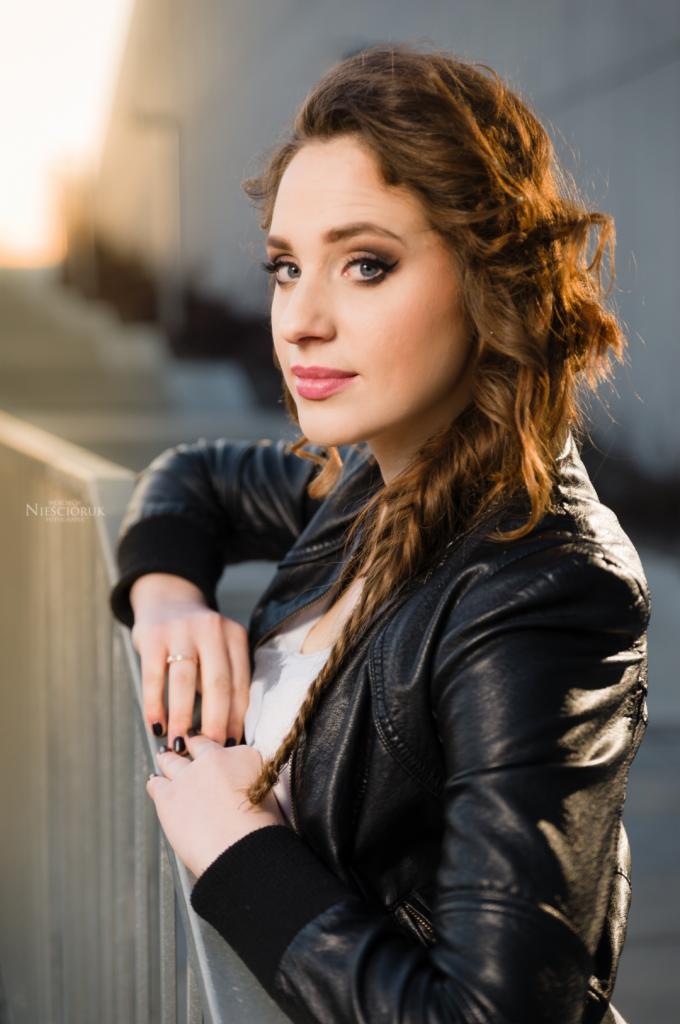 Portret młodej kobiety w skórzanej kurtce, opierającej się o metalową barierkę, z warkoczem i pierścionkiem na palcu, sfotografowana w ciepłym, zachodzącym słońcu.
