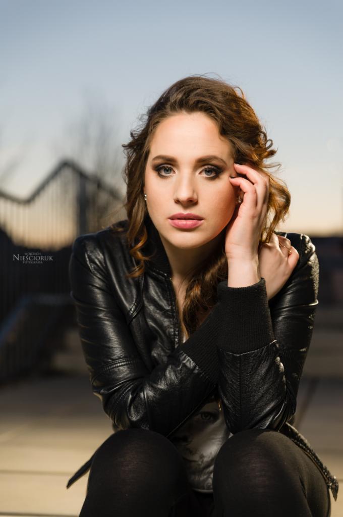 Portret młodej kobiety w czarnej skórzanej kurtce, z ręką przy twarzy, na tle miejskiego krajobrazu o zmierzchu.