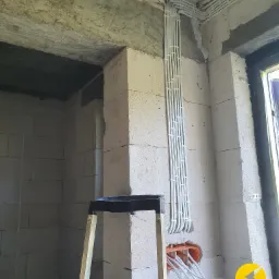 Białe kable elektryczne ułożone w peszlach prowadzone po surowych ścianach z betonu komórkowego, z widoczną puszką instalacyjną i wystającymi przewodami, fragment drabiny z boku.