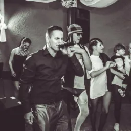 Wokalista w czarnej koszuli śpiewa do mikrofonu na scenie podczas występu, w tle zespół i publiczność w czarno-białych strojach, oświetlenie sceniczne.