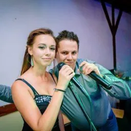 Duet wokalny: kobieta w czarnej sukience z cekinami i mężczyzna w szarej koszuli, oboje trzymają mikrofony i pozują, kobieta pokazuje znak zwycięstwa.