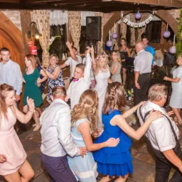 Wnętrze sali weselnej z parkietem, na kt&oacute;rym tańczą goście weselni, w tle widoczny zesp&oacute;ł muzyczny i dekoracje z kwiat&oacute;w i lampion&oacute;w.