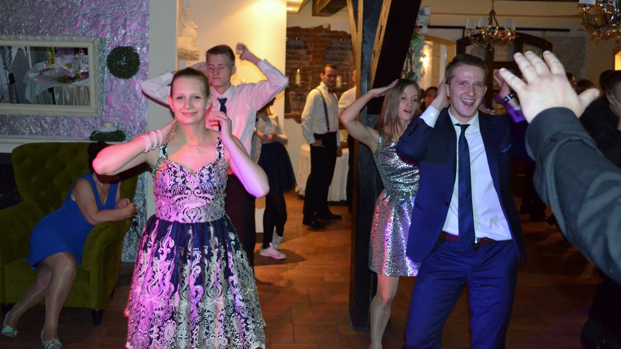 Uczestnicy zabawy tanecznej w eleganckich strojach, w tle dekoracje sali i siedząca osoba na zielonym fotelu.