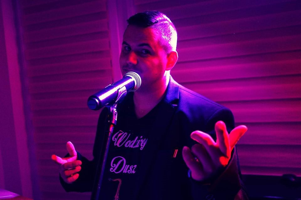 Mężczyzna w czarnej marynarce i koszulce z napisem 'Wodiry Dusz' śpiewa do mikrofonu oświetlonego fioletowym światłem.