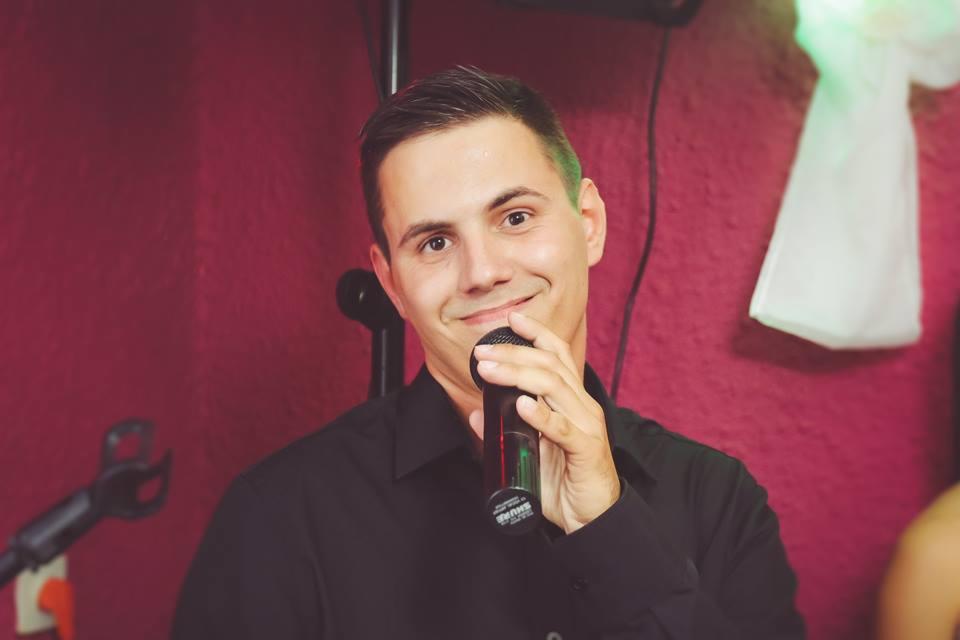 Uśmiechnięty mężczyzna w czarnej koszuli, trzymający mikrofon Shure na tle bordowej ściany z dekoracją.