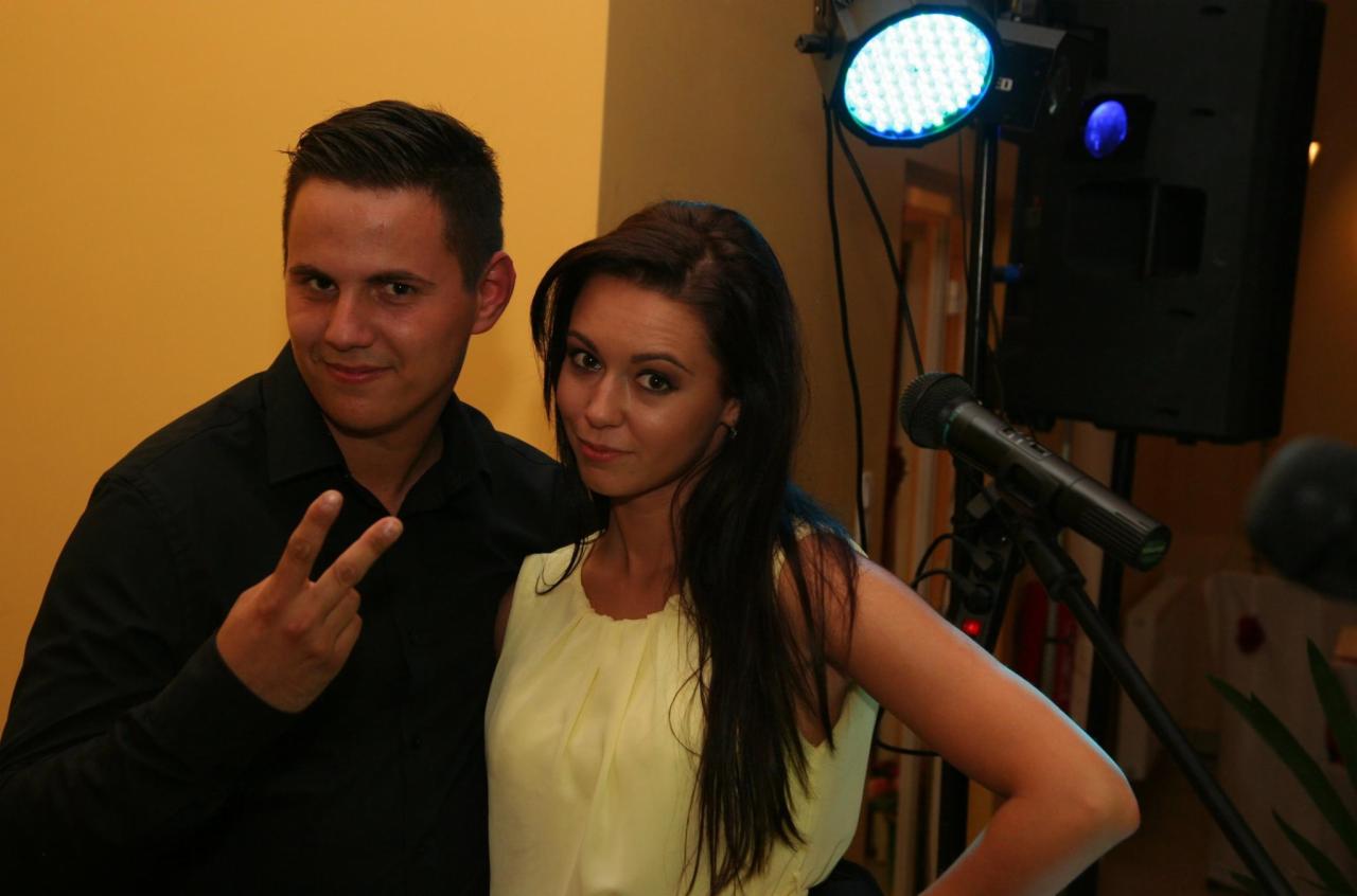 Para pozuje na tle sceny z mikrofonem i oświetleniem eventowym, mężczyzna pokazuje znak 'V'.