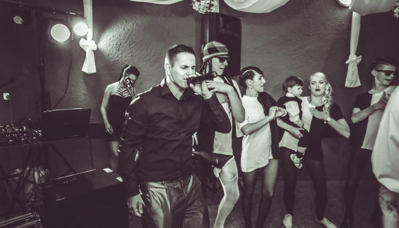 Wokalista w czarnej koszuli śpiewa do mikrofonu na scenie podczas występu, w tle zespół i publiczność w czarno-białych strojach, oświetlenie sceniczne.