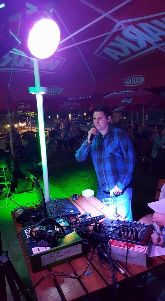 Mężczyzna w niebieskiej koszuli w kratę śpiewa do mikrofonu podczas karaoke na zewnątrz, z laptopem, mikserem dźwięku i oświetleniem scenicznym ustawionymi na drewnianym stole pod parasolami z logo...