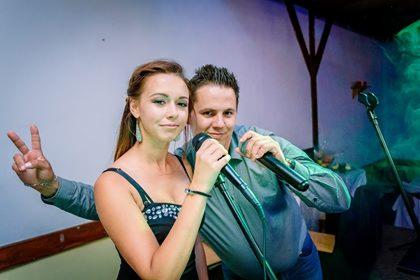 Duet wokalny: kobieta w czarnej sukience z cekinami i mężczyzna w szarej koszuli, oboje trzymają mikrofony i pozują, kobieta pokazuje znak zwycięstwa.