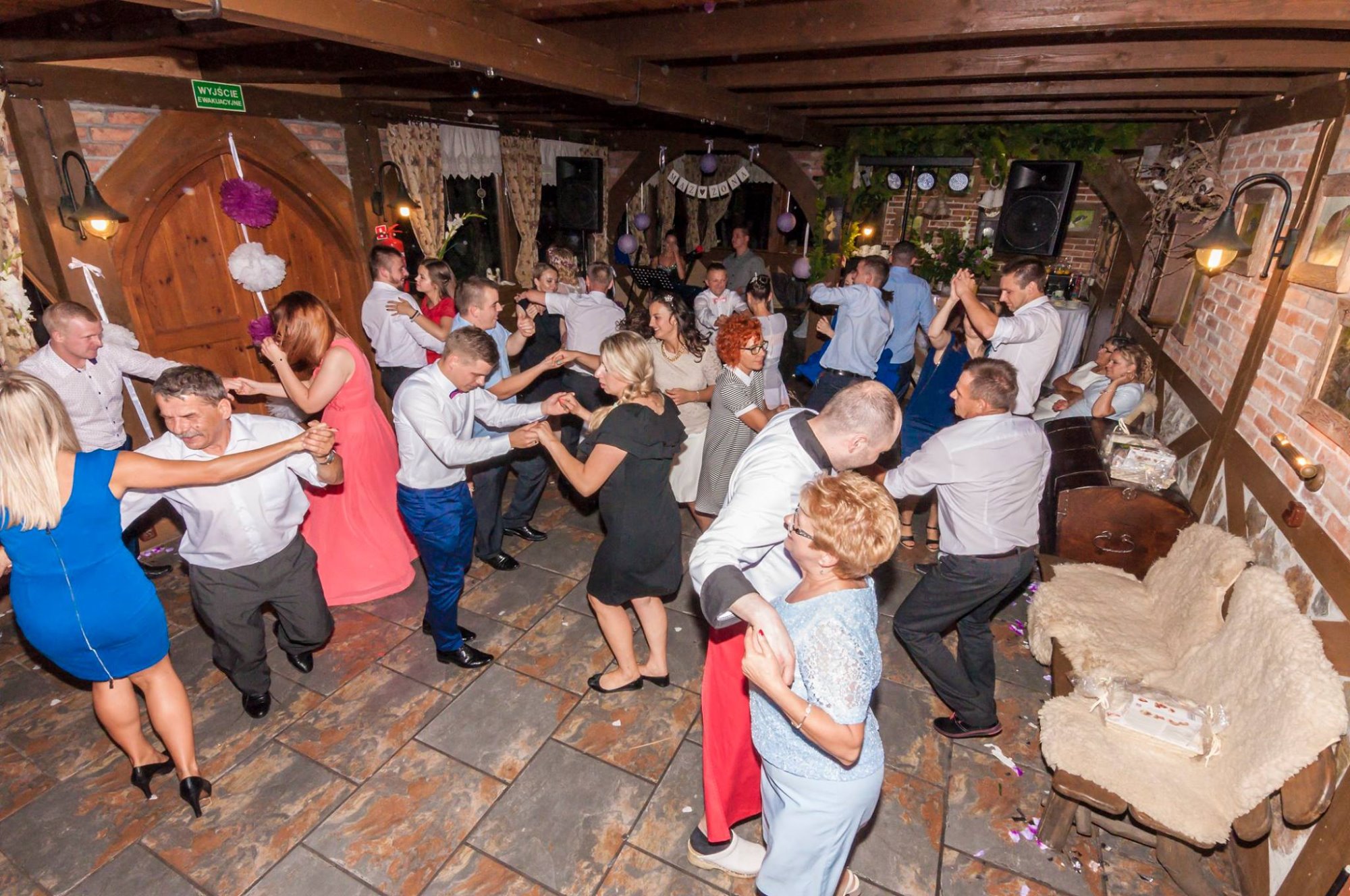 Goście weselni tańczą w rustykalnej sali z drewnianymi belkami, dekoracjami z kwiatów i balonów, oraz nagłośnieniem w rogu sali.