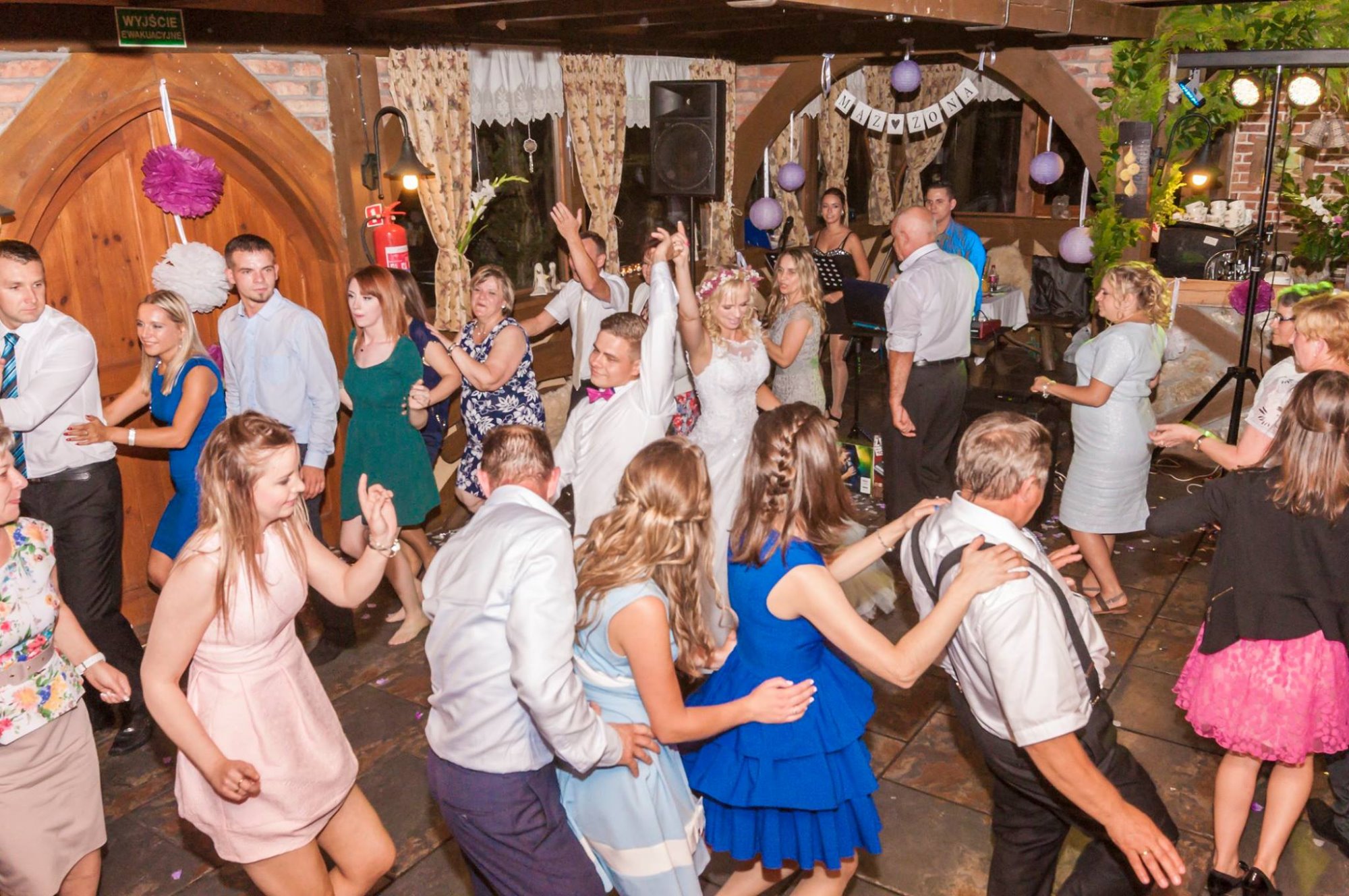 Wnętrze sali weselnej z parkietem, na którym tańczą goście weselni, w tle widoczny zespół muzyczny i dekoracje z kwiatów i lampionów.
