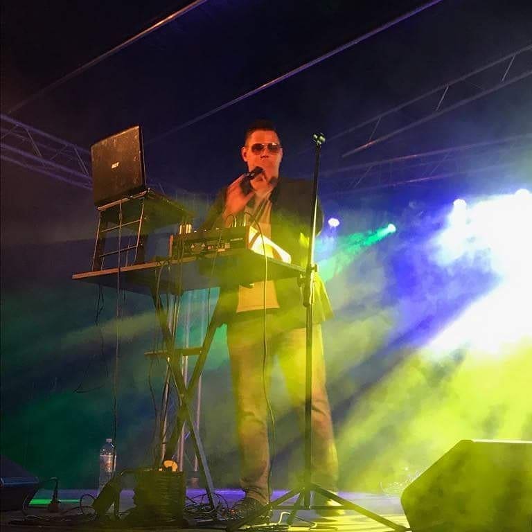 Mężczyzna w okularach śpiewa do mikrofonu na scenie oświetlonej zielonymi i żółtymi światłami, obok niego laptop na składanym stoliku.