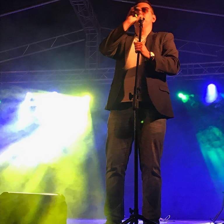 Wokalista w marynarce i okularach przeciwsłonecznych śpiewa do mikrofonu na scenie oświetlonej niebieskim i żółtym światłem.