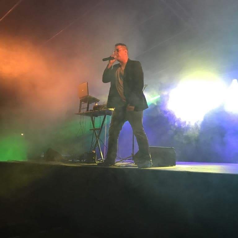 Wokalista w okularach, ubrany w marynarkę, śpiewa do mikrofonu na scenie oświetlonej kolorowym światłem i dymem, obok widoczny laptop na statywie.