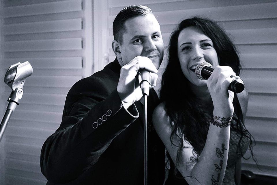 Duet wokalny w czerni i bieli: mężczyzna w garniturze i kobieta z tatuażem na ramieniu śpiewają do mikrofonów, trzeci mikrofon na statywie w tle.