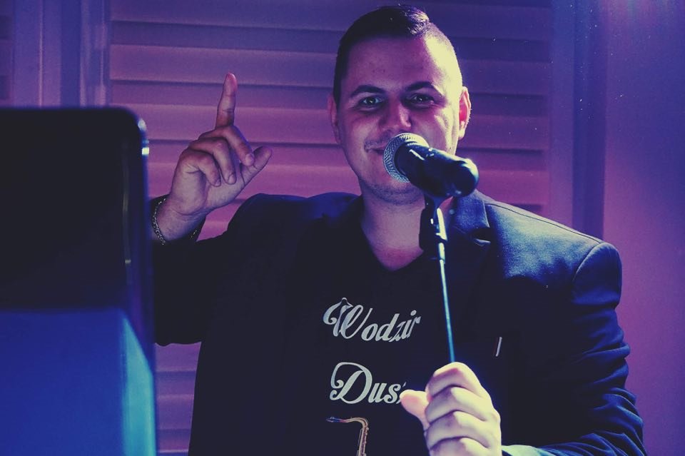 Mężczyzna z mikrofonem w ręku, w czarnej koszulce z napisem 'Wodzirej Dusz', wskazujący palcem do góry, oświetlony fioletowym światłem.