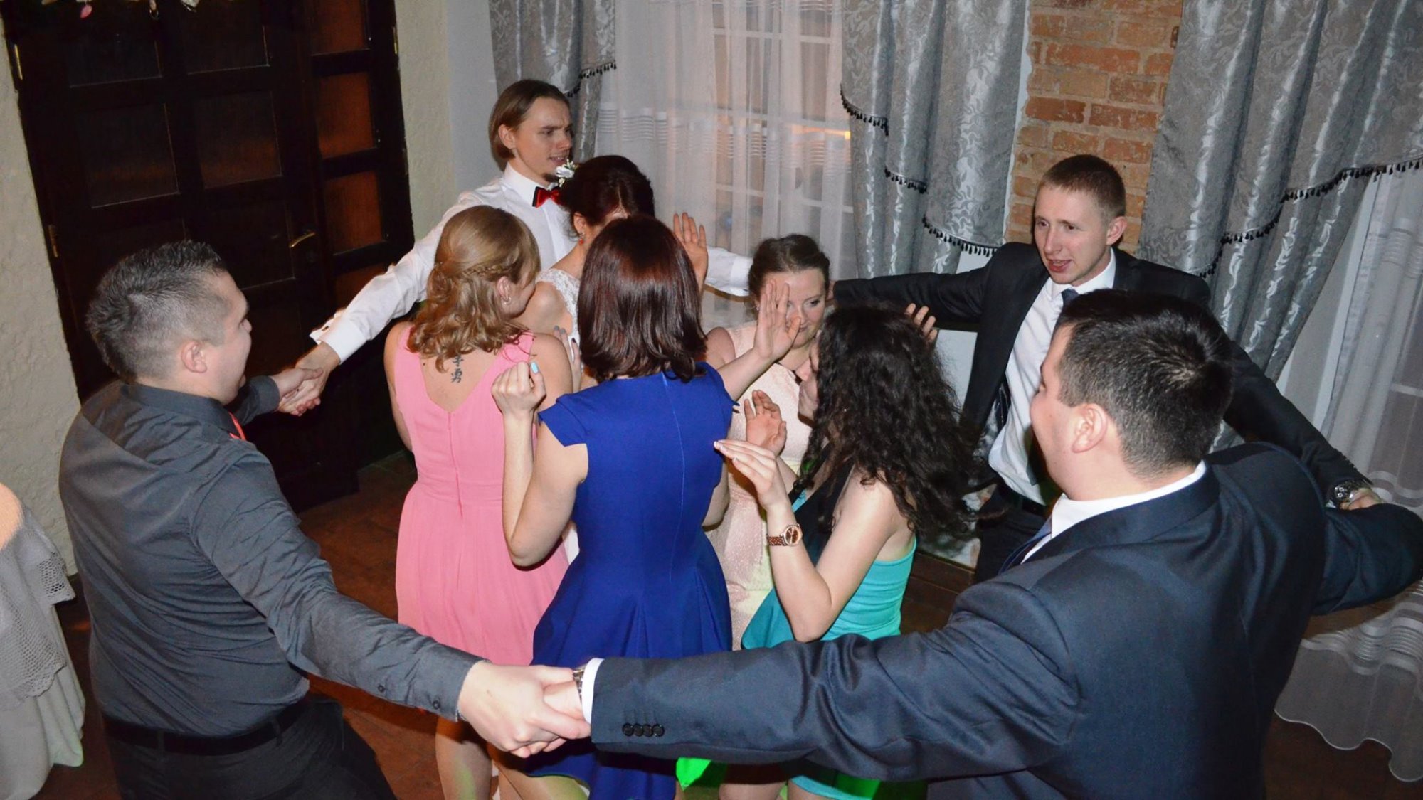 Goście w Rybniku tańczą w kręgu podczas uroczystości, widoczni mężczyźni w garniturach i kobiety w sukienkach, tło stanowią zasłony i ściana z cegły.