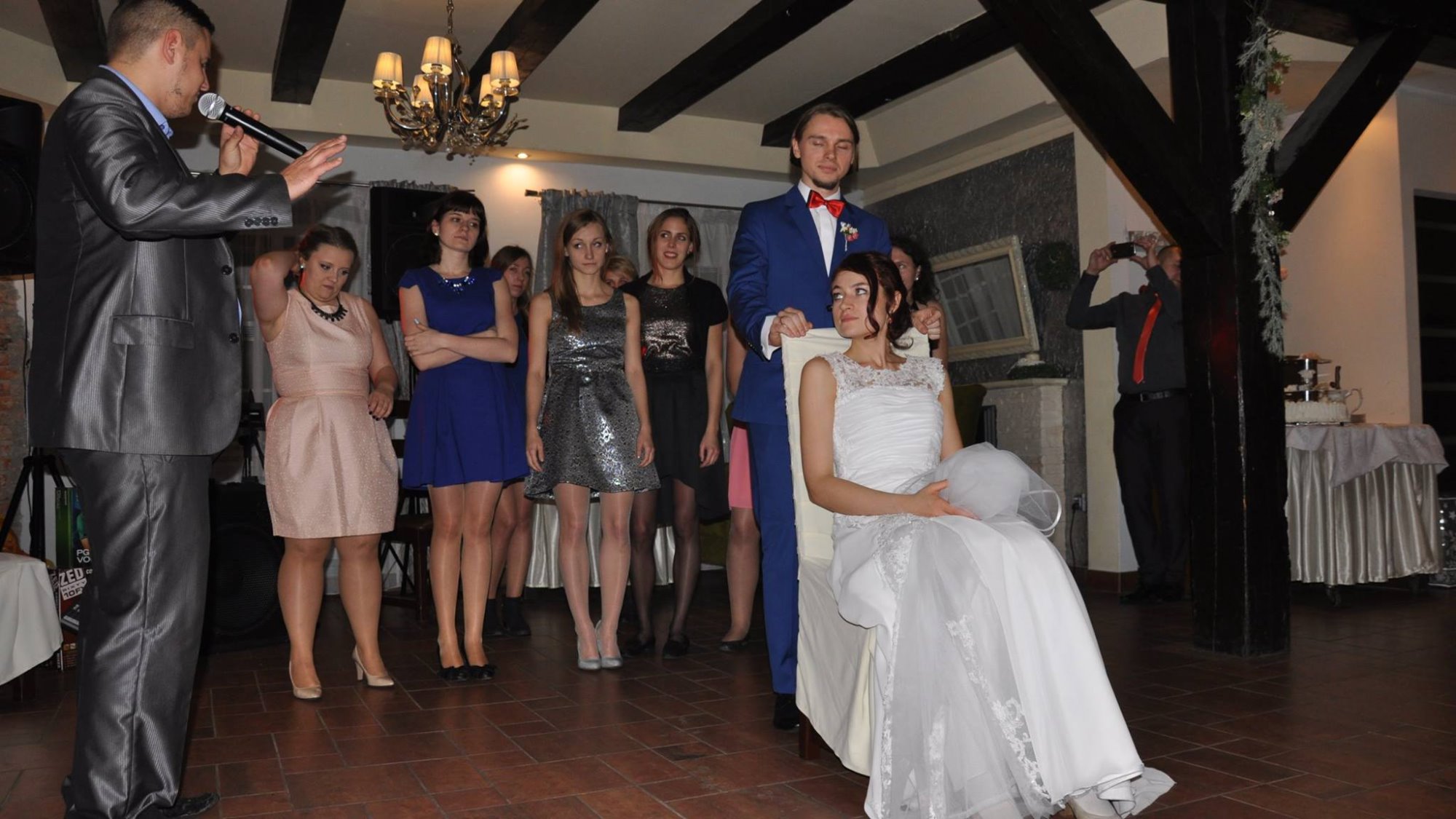 Oczepiny: Panna młoda siedzi na krześle, otoczona przez druhny i świadka w niebieskim garniturze, konferansjer z mikrofonem prowadzi zabawę, a fotograf robi zdjęcia w tle.