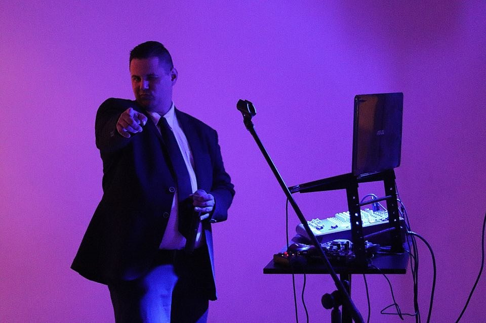 Mężczyzna w garniturze, wskazujący palcem w stronę widza, stoi obok mikrofonu i stolika z mikserem oraz laptopem ASUS w fioletowym oświetleniu scenicznym.