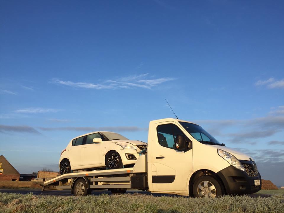 Biała laweta z białym samochodem osobowym na platformie, na tle błękitnego nieba z lekkimi chmurami i pola z niską trawą.