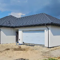 Niedawno ukończony parterowy dom szkieletowy z ciemną dachówką, jasną elewacją i bramą garażową, widoczny na tle błękitnego nieba z chmurami, na działce w trakcie prac ziemnych.