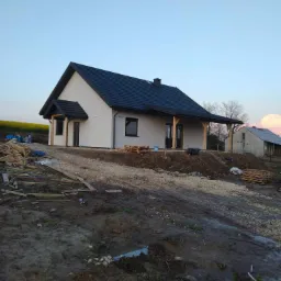 Niedokończony, parterowy budynek z ciemnym dachem i drewnianą konstrukcją werandy, otoczony rozkopanym terenem i stertą drewna, w tle pole rzepaku.