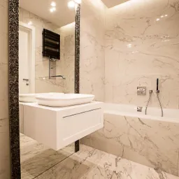 Nowoczesna łazienka z marmurowymi płytkami, wiszącą szafką z umywalką i dużym lustrem w ozdobnej ramie, oświetlona punktowymi lampami sufitowymi i listwą LED.