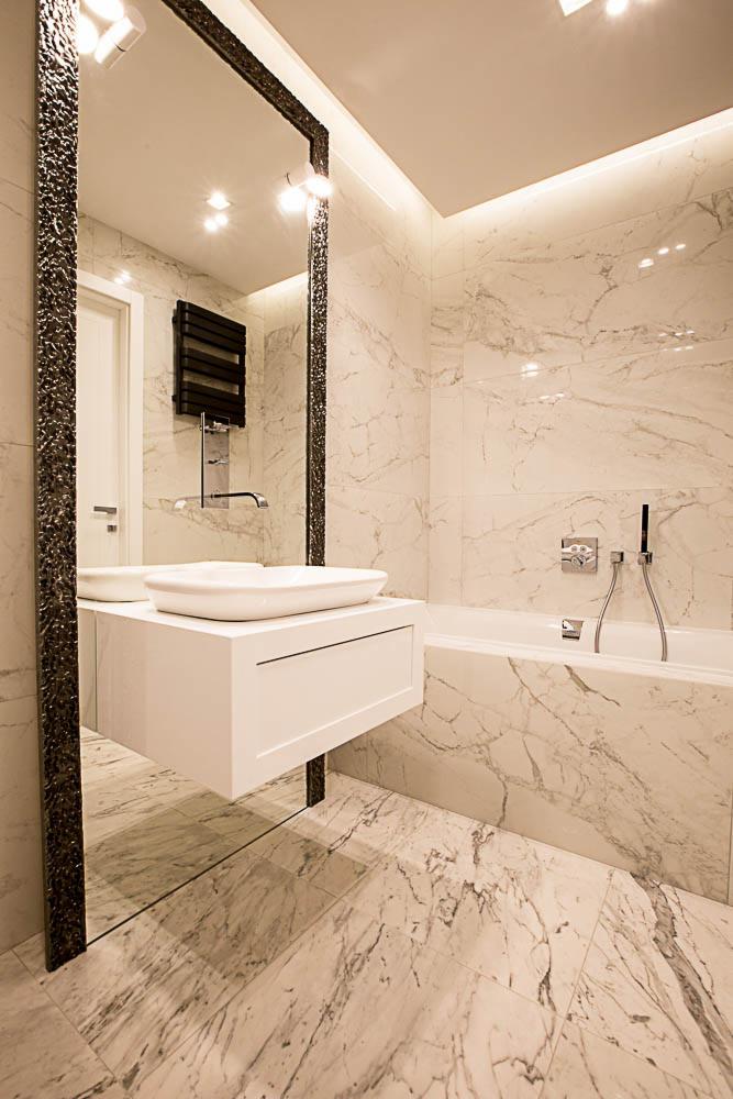 Nowoczesna łazienka z marmurowymi płytkami, wiszącą szafką z umywalką i dużym lustrem w ozdobnej ramie, oświetlona punktowymi lampami sufitowymi i listwą LED.